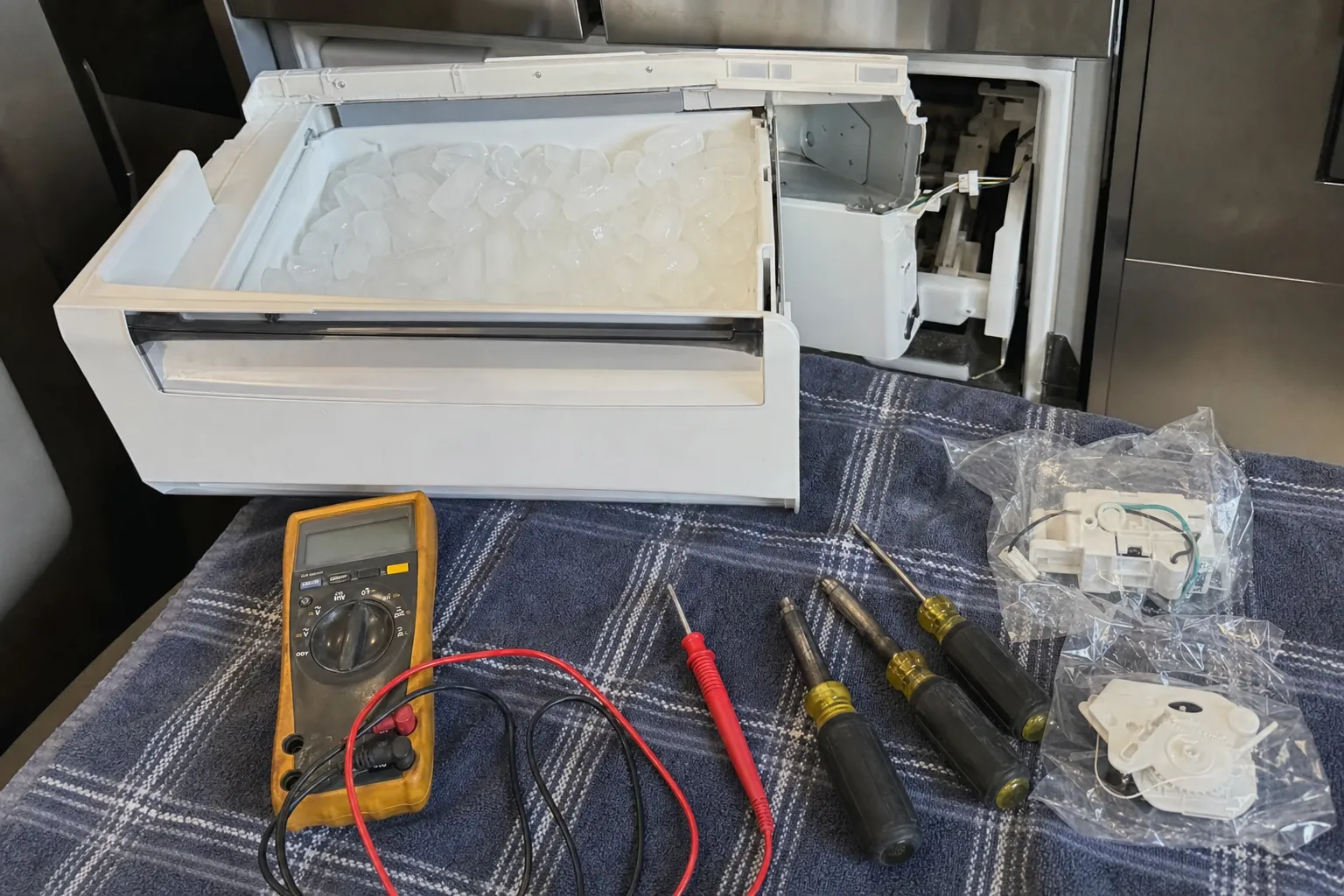 Ice drawer set on a towel beside a multimeter, screwdrivers and replacement ice maker parts still in their bags